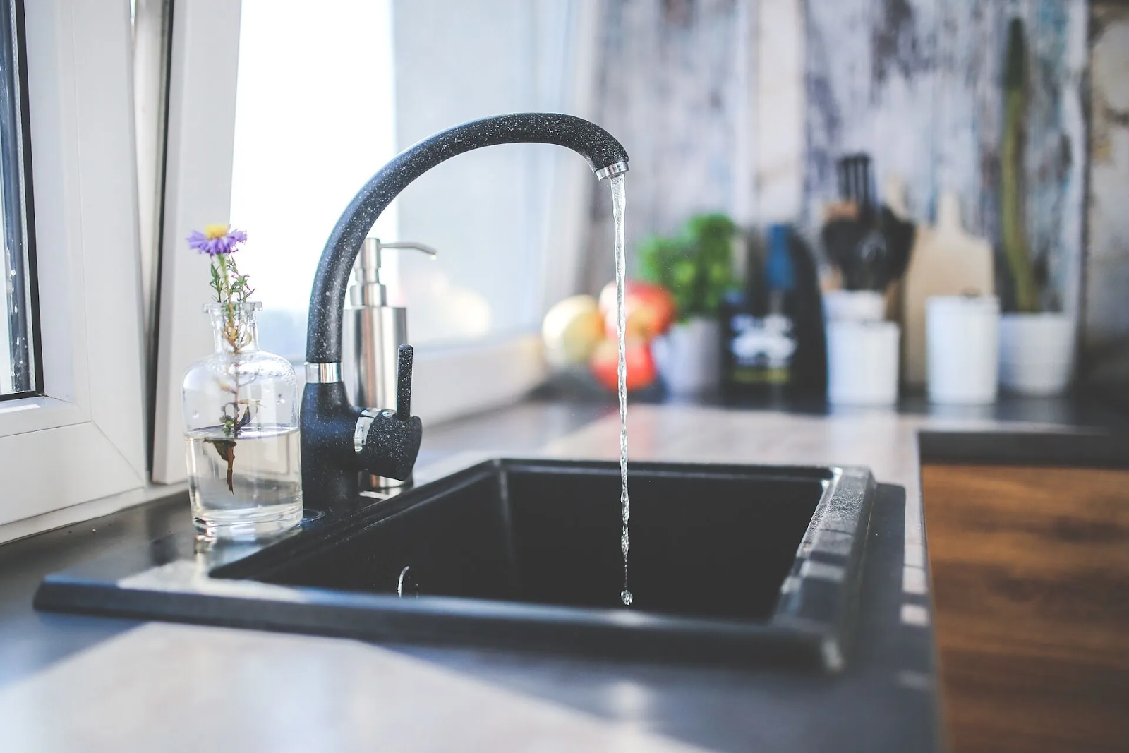 A kitchen sink running water with a small jar filled with water holding a single purple wildflower sitting beside it. A kitchen sink running water with a small jar filled with water holding a single purple wildflower sitting beside it.